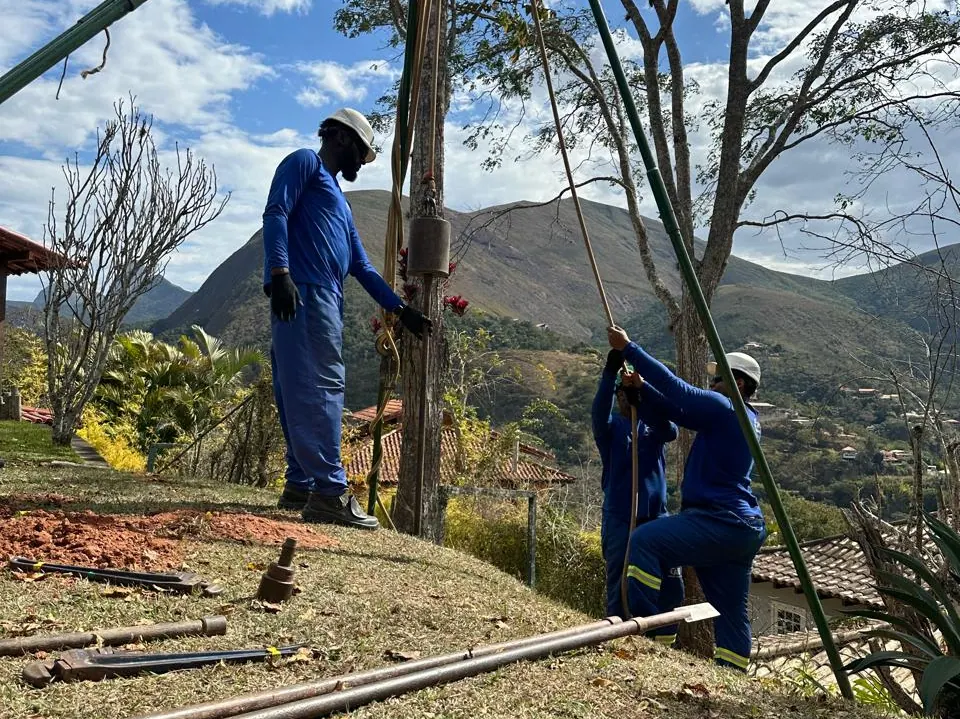 Equipe do Grupo CMG operando o tripé de sondagem em terreno de encosta com serra ao fundo