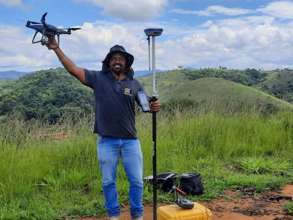 Profissional do Grupo CMG com drone em uma das mãos e o receptor GNSS na outra, em campo