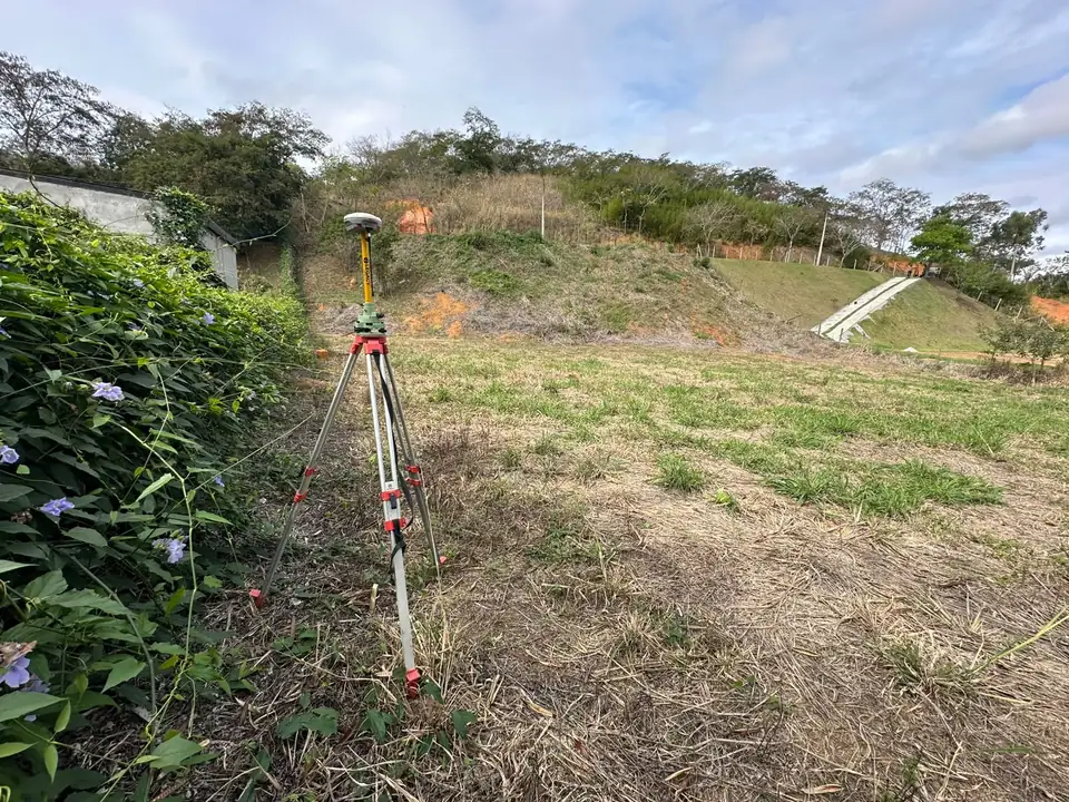 Receptor GNSS estacionado em tripé no lote já demarcado, com corte de terreno ao fundo